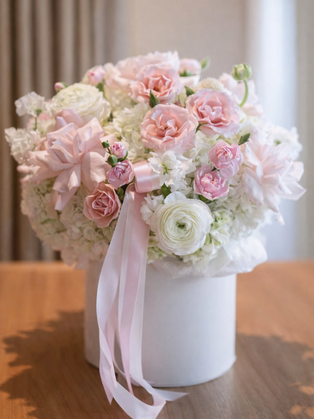Bouquet of pink and white flowers in a white flower box with a pink ribbon on a wooden surface.