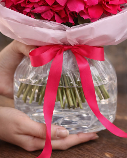 Bouquet of pink flowers in a aqua bag with a red ribbon held by a hand.
