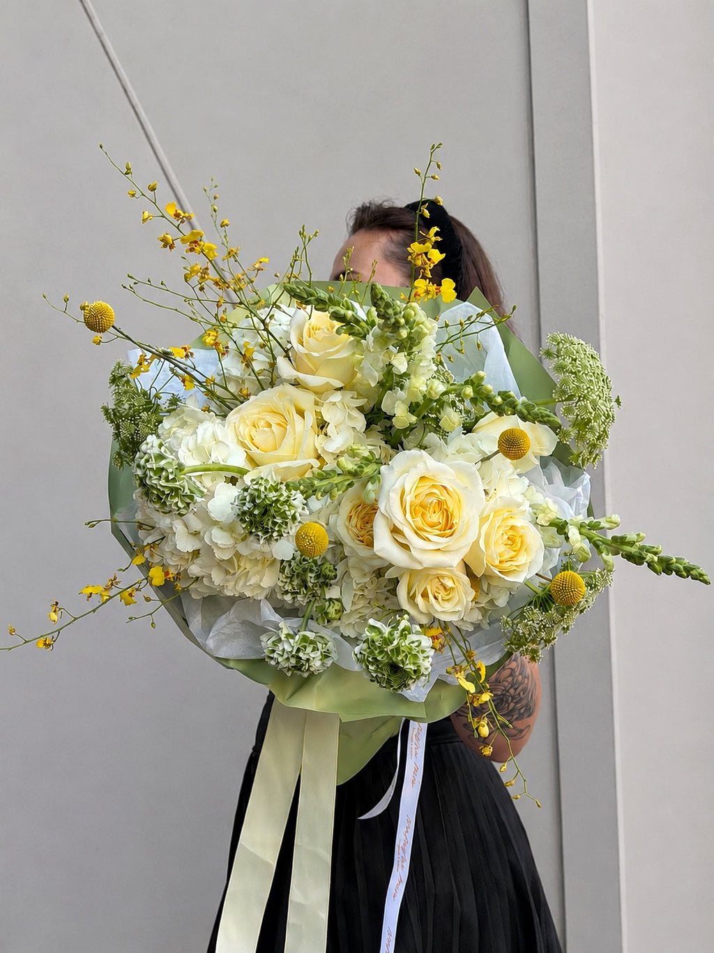 A large floral bouquet with pale yellow and white roses, green filler flowers, and small yellow blossoms, wrapped in pale green paper with cream and white ribbons. A person is partially visible behind the bouquet against a plain light gray wall.