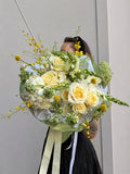 A large floral bouquet with pale yellow and white roses, green filler flowers, and small yellow blossoms, wrapped in pale green paper with cream and white ribbons. A person is partially visible behind the bouquet against a plain light gray wall.