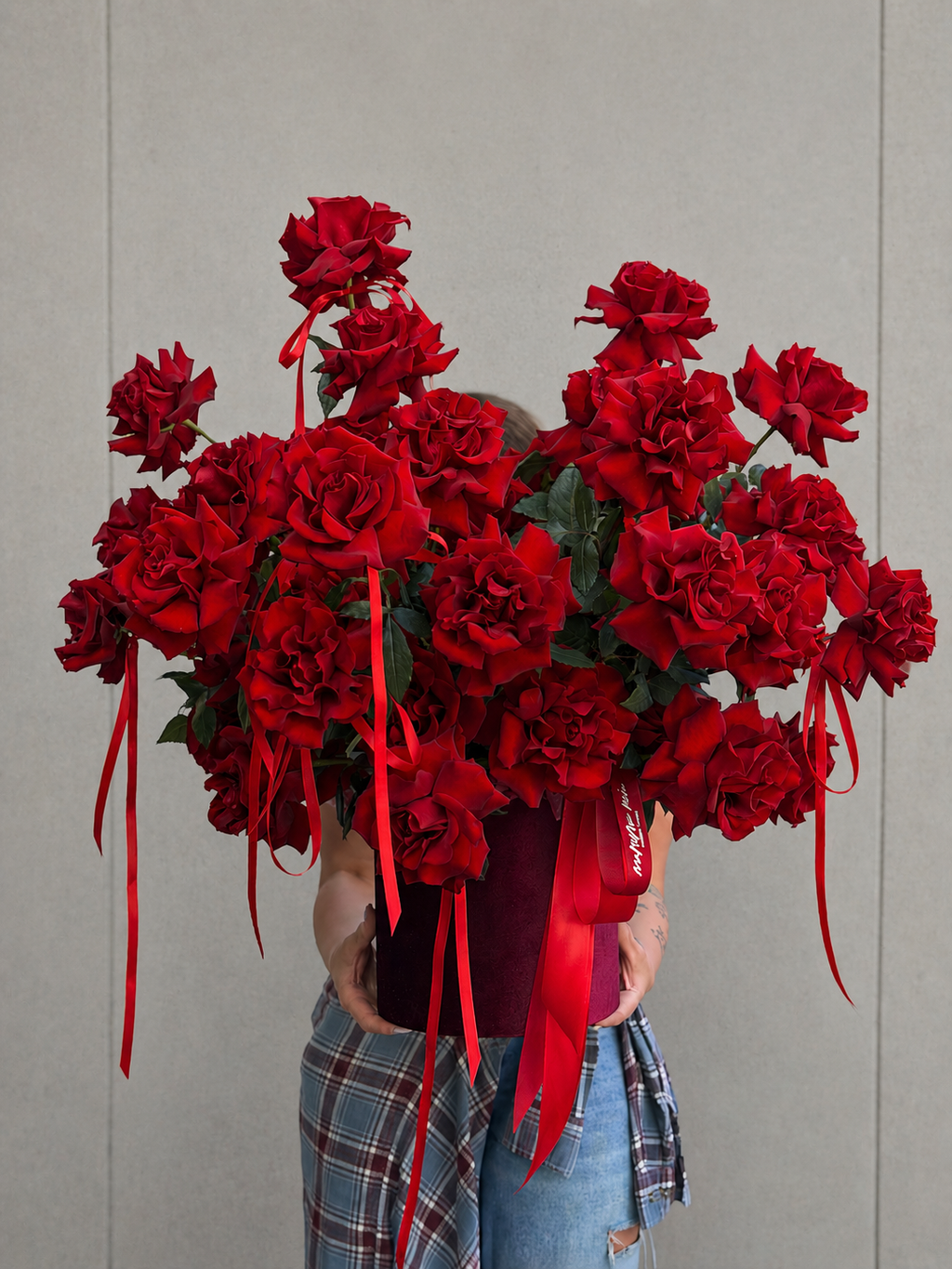 A large bouquet of deep red roses with long red ribbon ties, held by a person wearing a red top and blue plaid pants against a plain light gray wall.