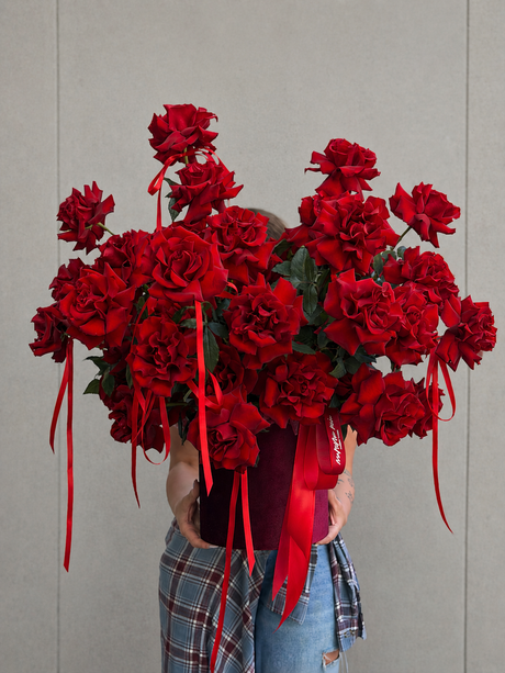 A large bouquet of deep red roses with long red ribbon ties, held by a person wearing a red top and blue plaid pants against a plain light gray wall.