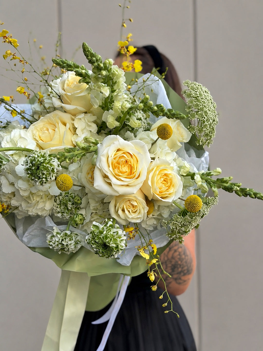 A large, vibrant bouquet of yellow and white flowers, including roses, hydrangeas, and green filler blooms, wrapped in pale green paper with a cream ribbon. A person is holding the bouquet in the background against a neutral beige wall.