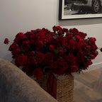 Bouquet of red roses in a woven basket on a couch with a framed picture in the background.