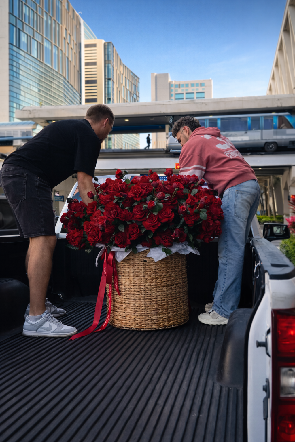 Two people loading a large basket of red flowers into the back of a pickup truck in an urban setting.