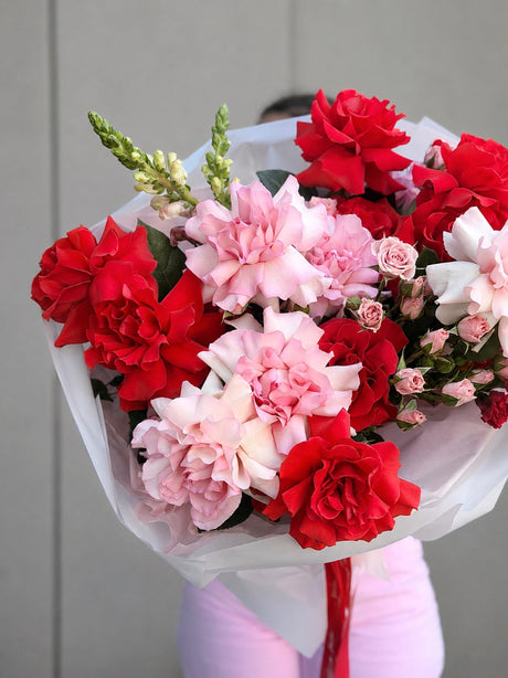 A vibrant bouquet of red and pink flowers wrapped in white tissue paper, held by a person wearing light pink pants against a neutral gray paneled wall.