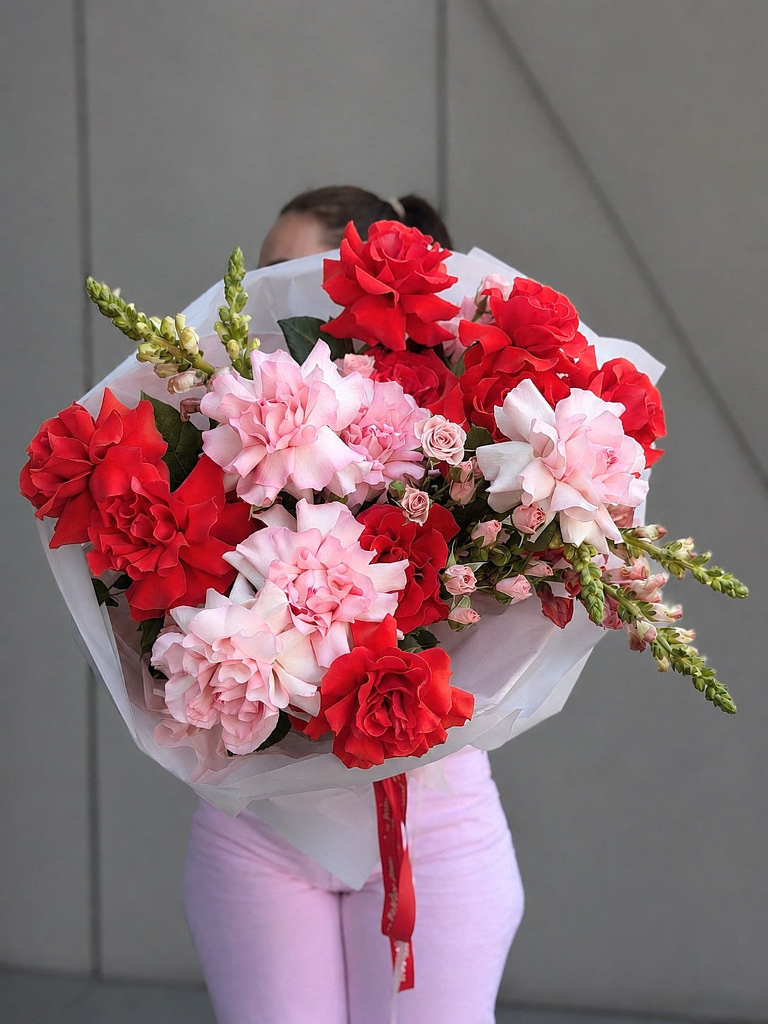 A person holding a large bouquet of red and pink roses flowers, including roses and carnations, wrapped in white tissue paper with a red ribbon, against a gray paneled wall.