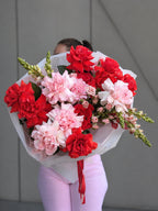 A person holding a large bouquet of red and pink roses flowers, including roses and carnations, wrapped in white tissue paper with a red ribbon, against a gray paneled wall.