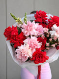 A vibrant bouquet of red and pink flowers wrapped in white tissue paper, held by a person wearing light pink pants against a neutral gray paneled wall.
