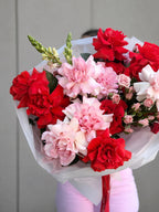 A vibrant bouquet of red and pink flowers wrapped in white tissue paper, held by a person wearing light pink pants against a neutral gray paneled wall.