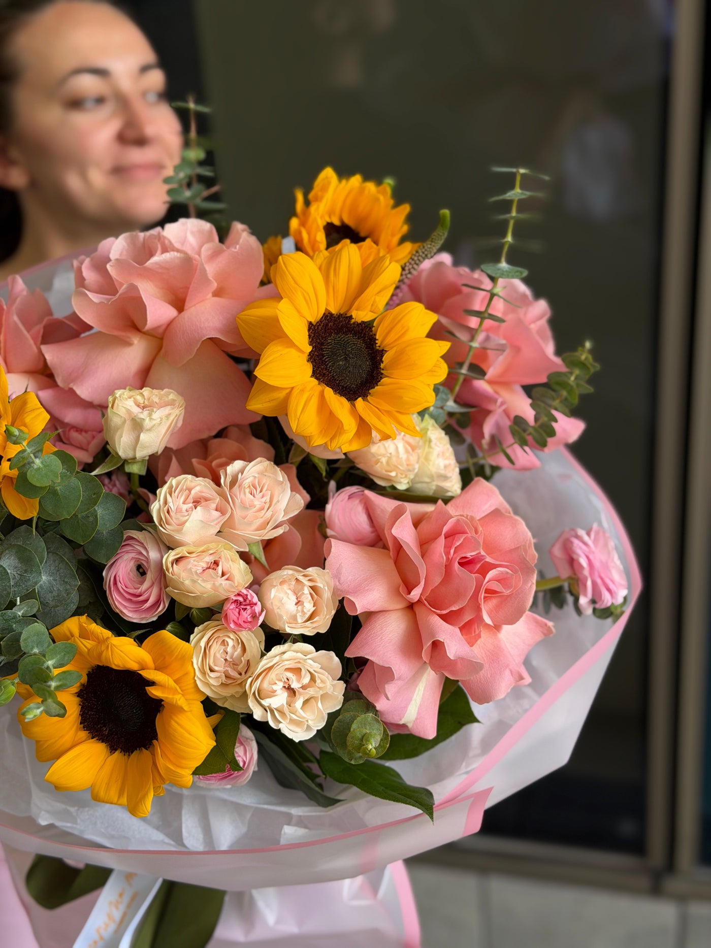 A vibrant floral bouquet featuring yellow sunflowers, pink and peach roses, and green eucalyptus stems, wrapped in white paper with pink accents. A woman is softly blurred in the background.