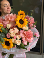 A vibrant floral bouquet featuring yellow sunflowers, pink and peach roses, and green eucalyptus stems, wrapped in white paper with pink accents. A woman is softly blurred in the background.